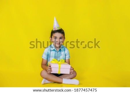 A little boy sits on the floor and holds a gift on a yellow background.A birthday present.the concept of celebration and fun.a cap on the boy's head.