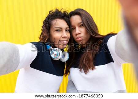 Two funny Hispanic girls taking selfie photo on mobile phone. They are isolated on a yellow background. Concept of friendship.