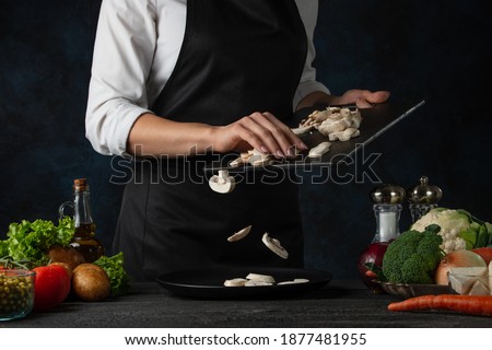 The professional chef in black apron pours chopped mushrooms on the plate for cooking soup on dark blue background. Backstage of preparing meal. Healthy dish. Food concept. Frozen motion. Cookbook.