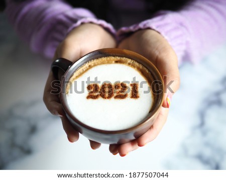 Number 2021 on frothy surface of cappuccino served in coffee cup holding by female hands with acrylic paint nail polish. Holidays food art theme for New Year 2021 celebration. (selective focus)
