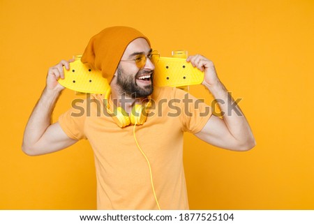 Funny cheerful laughing young bearded man 20s wearing basic casual t-shirt headphones eyeglasses hat standing hold skateboard looking aside isolated on bright yellow colour background studio portrait
