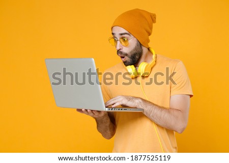 Shocked amazed young bearded man 20s wearing basic casual t-shirt headphones eyeglasses hat standing hold working on laptop pc computer isolated on bright yellow colour background, studio portrait