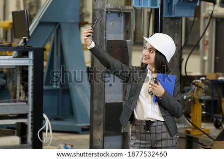 Young female businesswoman in a business suit and black glasses with a blue notebook and smartphone making selfy, controlling work process in a factory. 