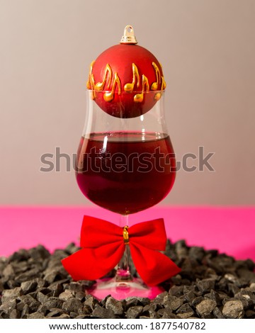 Glass of wine decorated with glass ball ornament and red bow, placed in a center of decorative pebbles. Grey and pink background. Holiday concept.