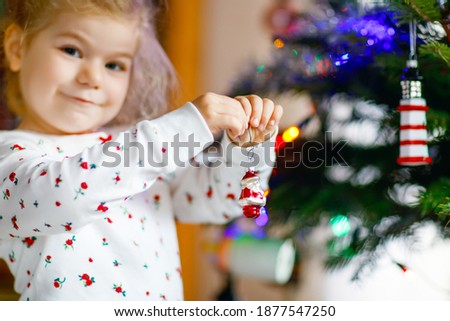 Adorable toddler girl in pajamas decorating Christmas tree with toy in cute hands. Little child in nightwear standing by Xmas tree. celebration of traditional family winter holiday