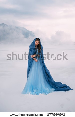 Art photo. Fantasy young woman fairy elf in blue cape with hood stands in cold wind. Winter nature background, mountains in the clouds, dramatic sky white snow. Girl Queen walks in dress, silk cloak.