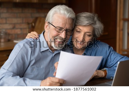 Happy bonding old senior family couple looking through paper correspondence, reading letter with good news. Curious middle aged spouses checking bills or medical insurance contract together at home. Royalty-Free Stock Photo #1877561002
