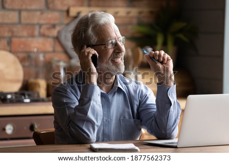 Happy middle aged mature businessman in eyeglasses holding mobile phone talk distracted from computer work. Smiling senior man enjoying cellphone conversation at home office, multitasking concept.