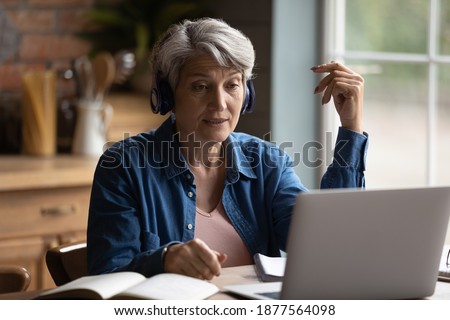 Focused middle aged mature woman wearing wireless headphones, involved in online communication with teacher using computer software video call application, older people and distant education concept. Royalty-Free Stock Photo #1877564098