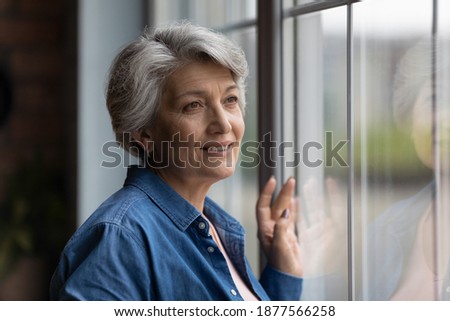 Dreamy smiling elderly 60s hoary woman looking in distance out of window, recollecting good memories or visualizing future at home, planning vacation or enjoying peaceful mindful day alone indoors. Royalty-Free Stock Photo #1877566258
