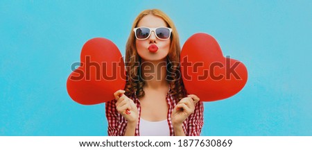 Portrait of happy young woman holding red heart shaped balloon and blowing lips sending sweet air kiss on a blue background