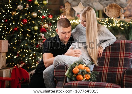 Adorable marriage couple sit near Christmas tree and fireplace at home, caring husband is stroking the stomach beautiful pregnant wife, young family enjoy happiness, winter holidays concept