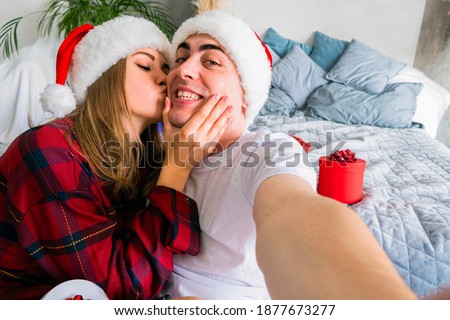 Online Christmas Party. Woman kissing man taking selfie wearing santa hats and red pajamas against the background of New Year's gifts