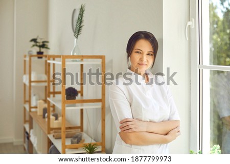 Portrait of serious cosmetologist or aesthetic nurse in her workplace. Professional beautician with clipboard standing in beauty salon. Female massagist in massage room looking at camera Royalty-Free Stock Photo #1877799973