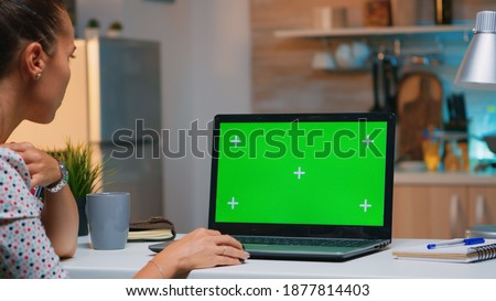 Freelancer looking at green screen monitor sitting at home in kitchen. Business woman watching desktop laptop display with green mockup, chroma key, during night time working overtime.