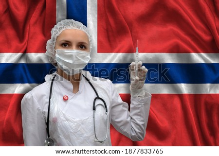 Young woman doctor in medical uniform on the background of the national flag of Norway is holding a syringe. The concept of a new vaccine against the disease, flu vaccine vaccination