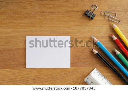 On a wooden table lay down a white card, multi colored pencils, clips and a linear, table top with space for text