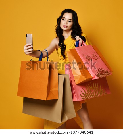 Young brunette woman in stylish yellow dress online shopping with modern pink orange and brown shopping bags makes selfie using her mobile smartphone on yellow background. Big Sales