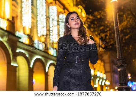 Christmas shooting, a pretty brunette girl in the city at night next to the Christmas lights of a beautiful building. Elegant black christmas or new year's eve dress with black boots