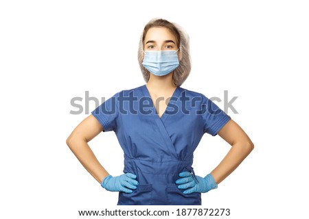 Young beautiful female doctor in a blue surgical gown, blue glove, a surgeon's cap and a medical mask on her face holds her hands on a belt, isolated on a white background. Copspace