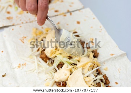 The girl carefully pours mayonnaise from a spoon onto an almost finished shawarma.