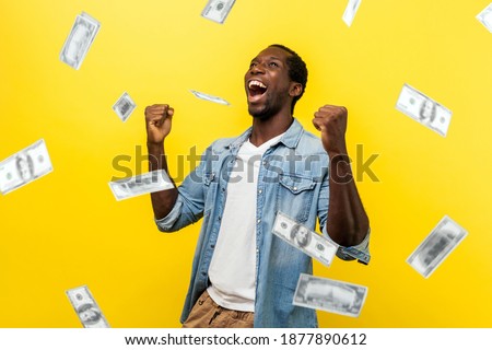 Money rain, winner and rich. happy young ecstatic motivated man standing with raised fists and shouting for joy, winner excited for success. dollars falling. indoor isolated on yellow background