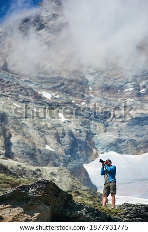 Male tourist using camera to take photo of beautiful mountains scenery on background. Mountain hiking, man reaching peak in sunny summer day. Concept of travelling and professional photography.