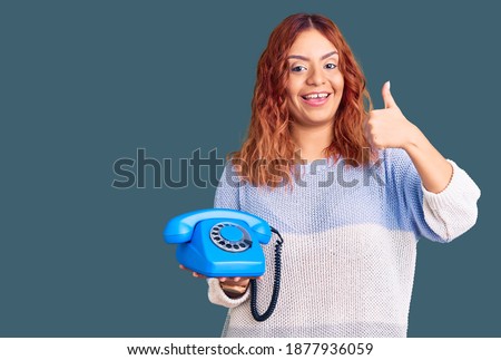 Young latin woman holding vintage telephone smiling happy and positive, thumb up doing excellent and approval sign 