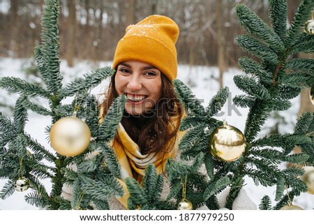 Happy smiling girl outside at winter behind synthetic fir tree decorated with shiny christmas balls. She wears yellow knit scarf and hat.