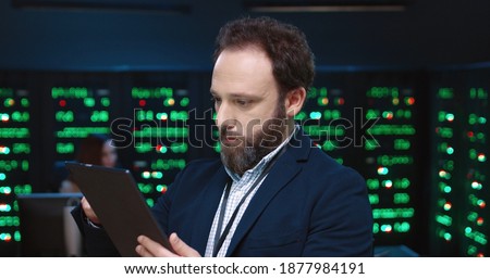 Close up of system administrator programmer in jacket working on tablet standing in office on background of working servers.