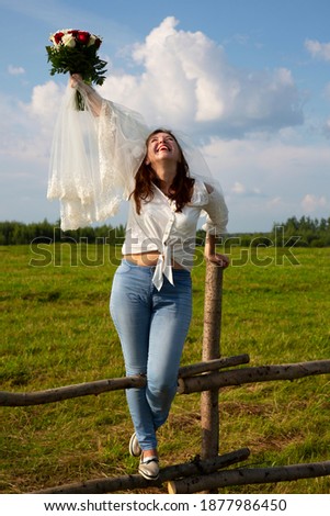 Brown-haired woman in a veil and jeans with a bouquet of red and white roses on the background of a rural field and the sky, a bride's bouquet, a budget modern wedding