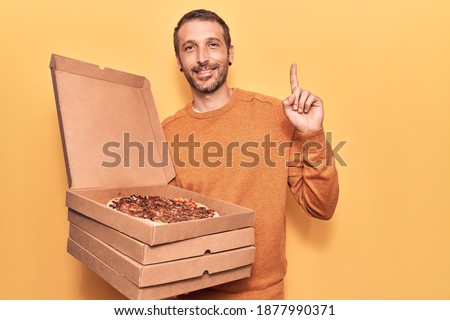 Young handsome man holding delivery pizza boxes smiling with an idea or question pointing finger with happy face, number one 