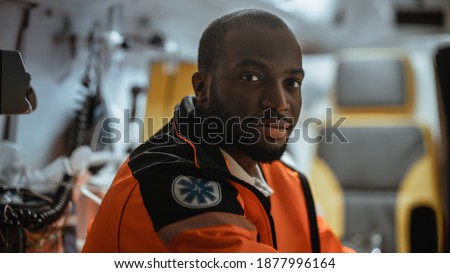 Black African American Paramedic Looking into Camera in an Ambulance Vehicle Going for Emergency. Emergency Medical Technicians are on Their Way to a Call Outside the Healthcare Hospital. Royalty-Free Stock Photo #1877996164