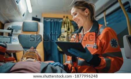 Female EMS Professional Paramedic Using Tablet Computer to Fill a Questionnaire for the Injured Patient on the Way to Hospital. Emergency Care Assistant Comforting the Patient in an Ambulance. Royalty-Free Stock Photo #1877996248