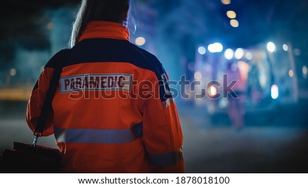 Female EMS Paramedic Proudly Standing With Her Back Turned to Camera in High Visibility Medical Orange Uniform with "Paramedic" Text Logo. Successful Emergency Medical Technician or Doctor. Royalty-Free Stock Photo #1878018100