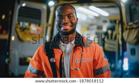 Portrait of a Black African American EMS Paramedic Proudly Standing in Front of Camera in High Visibility Medical Orange Uniform and Smiling. Successful Emergency Medical Technician or Doctor at Work. Royalty-Free Stock Photo #1878018112