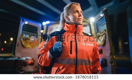 Portrait of a Female EMS Paramedic Proudly Standing in Front of Camera in High Visibility Medical Orange Uniform with "Paramedic" Text Logo. Successful Emergency Medical Technician or Doctor at Work. Royalty-Free Stock Photo #1878018115