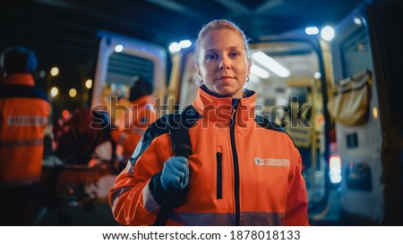 Portrait of a Female EMS Paramedic Proudly Standing in Front of Camera in High Visibility Medical Orange Uniform with "Paramedic" Text Logo. Successful Emergency Medical Technician or Doctor at Work. Royalty-Free Stock Photo #1878018133