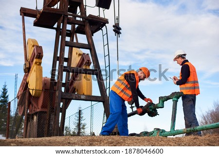 Full length view of two oil workers wearing overalls, orange vests, and helmets, working on oil field next to oil pump jack, using industrial wrench, tightening bolt on pipe, the other making notes