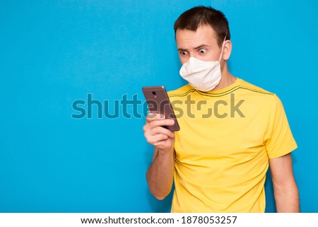 Portrait anxious young man in medical mask against virus looking at phone seeing bad news or photos with disgusting emotion on face isolated on blue background. Human emotion, reaction, expression