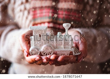 Women's hands in a knitted winter white sweater and holding a Christmas decor in the form of a silver steam locomotive. Added the effect of falling snow on the photo.