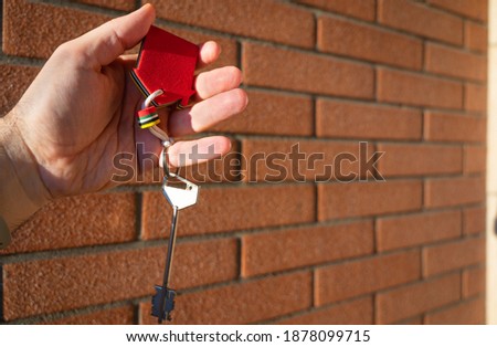 Home buying conceptual image: open hand offers big key with red house silhouette keychain. In the background a red brick wall.