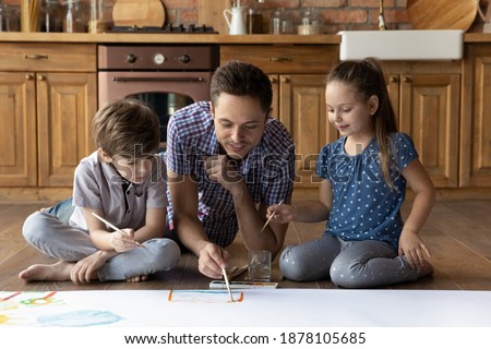 Study in play. Caring single father drawing big picture with son daughter kids on heating floor. Two junior children sister brother learn to paint using brush watercolor watching young man art teacher