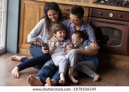 Kidding, giggling, having fun. Happy cute family of four cuddle on warm heated floor at kitchen. Millennial mommy daddy little kids daughter son laughing tickling messing shooting funny video on phone