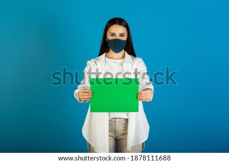 Young woman in white shirt and medical mask is holding green sheet of paper isolated on blue background. Copy space.