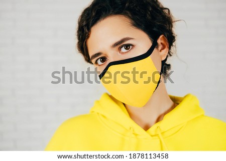 Female in bright yellow mask and hoodie with curly hair looking at camera during pandemic