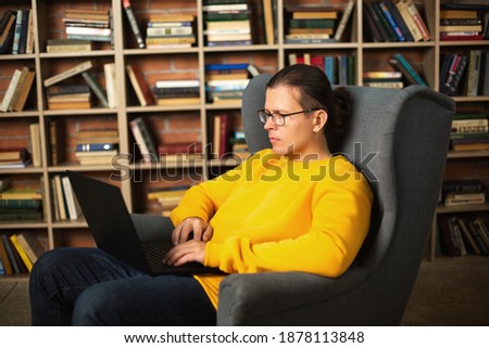 Smart adult male in casual clothes and glasses resting in comfortable armchair and browsing laptop against bookshelf in cozy library