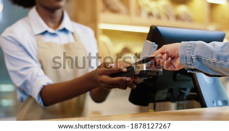 Close up of African American female seller in mask and protective gloves selling food in bakery shop. Client hand paying with credit card on device. Small business. Shopping. Bakehouse concept