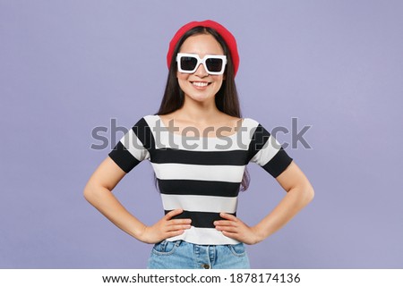 Smiling young brunette asian woman 20s wearing striped t-shirt red beret dark eyeglasses standing with arms akimbo on waist looking camera isolated on pastel violet colour background studio portrait