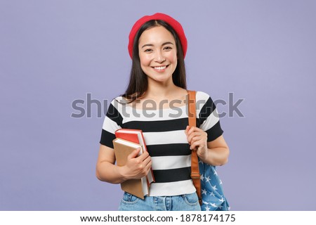Smiling attractive young brunette asian student woman wearing striped t-shirt red beret backpack standing hold in hand books looking camera isolated on pastel violet colour background studio portrait